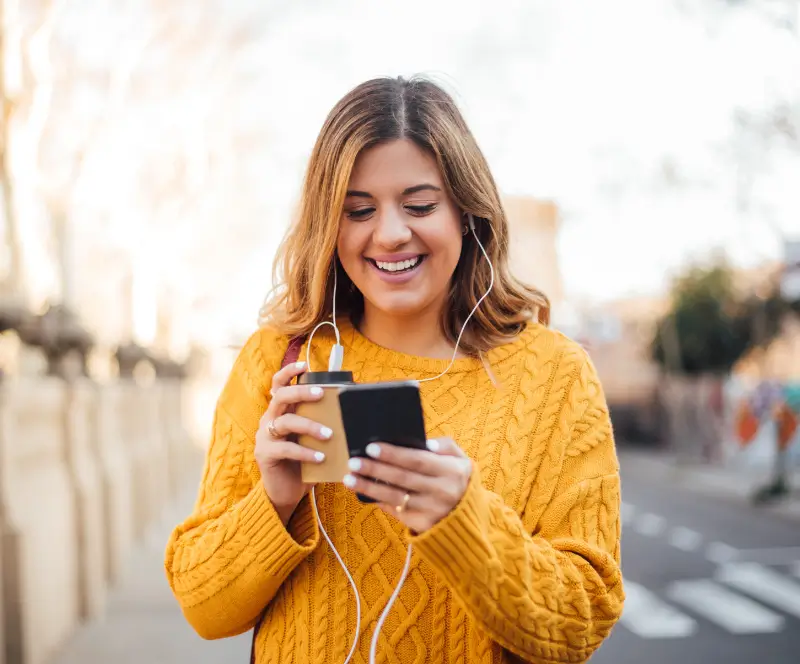 A woman walking down the street with headphones laughing at her phone